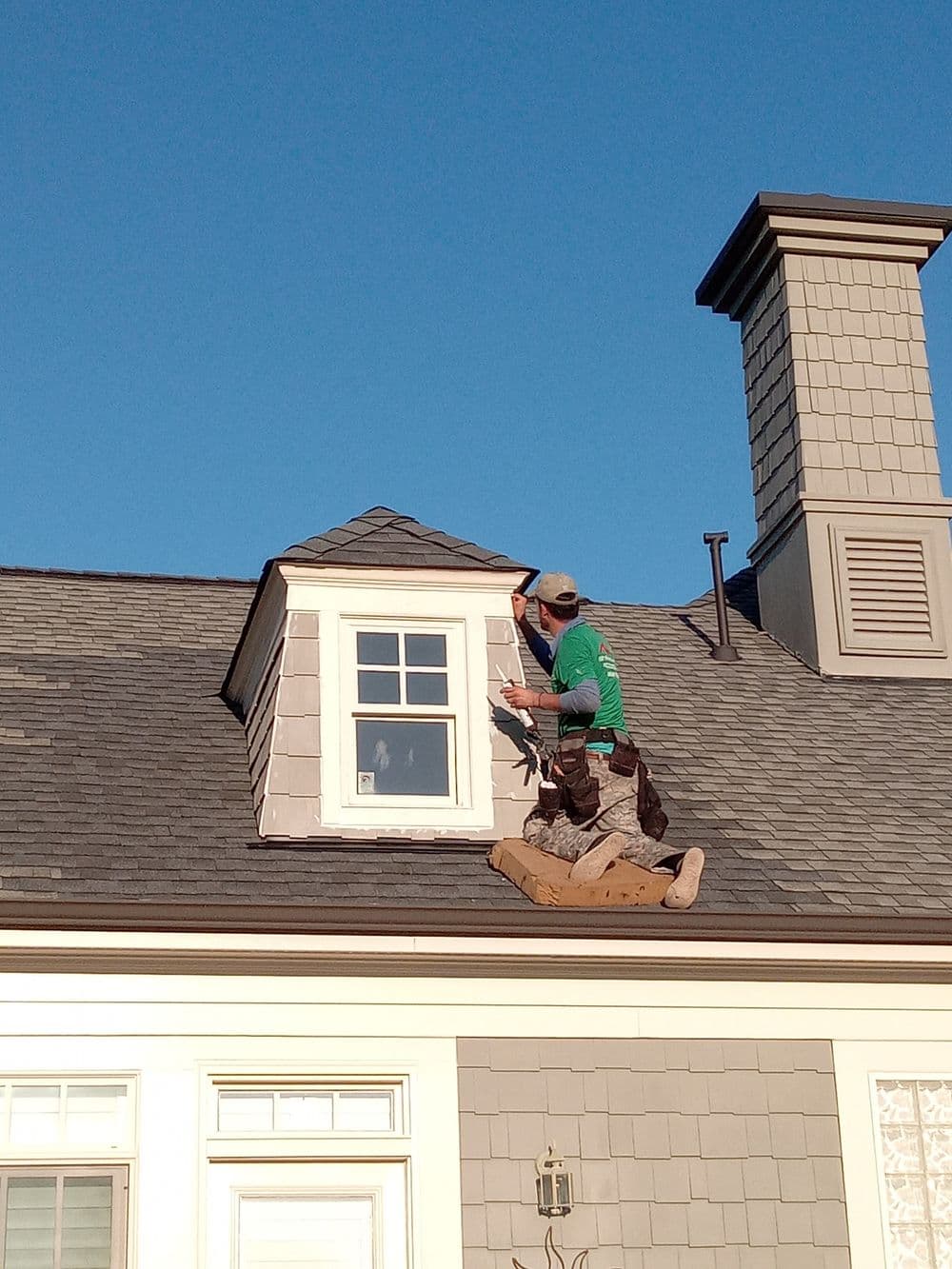 Roofer working on a gable roof, repairing window trim under clear blue sky.