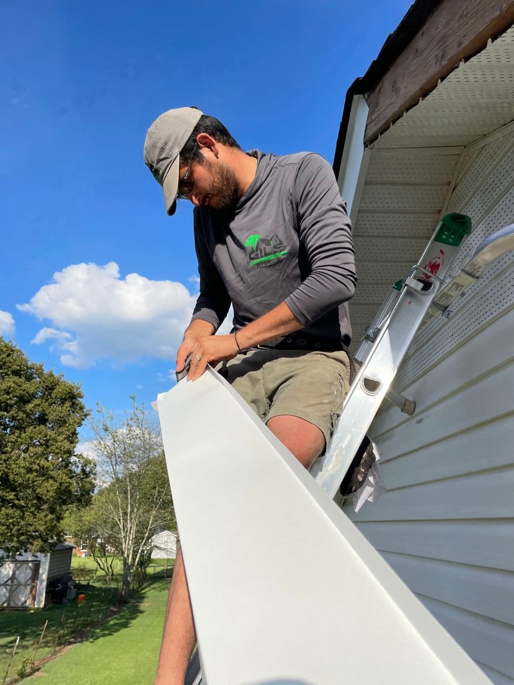 Man installing roof trim while standing on a ladder, under a blue sky with clouds.