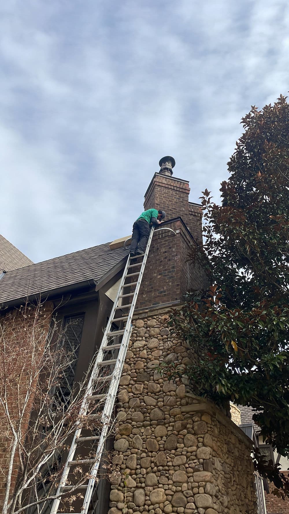Worker on ladder inspecting chimney of a residential home under a cloudy sky.