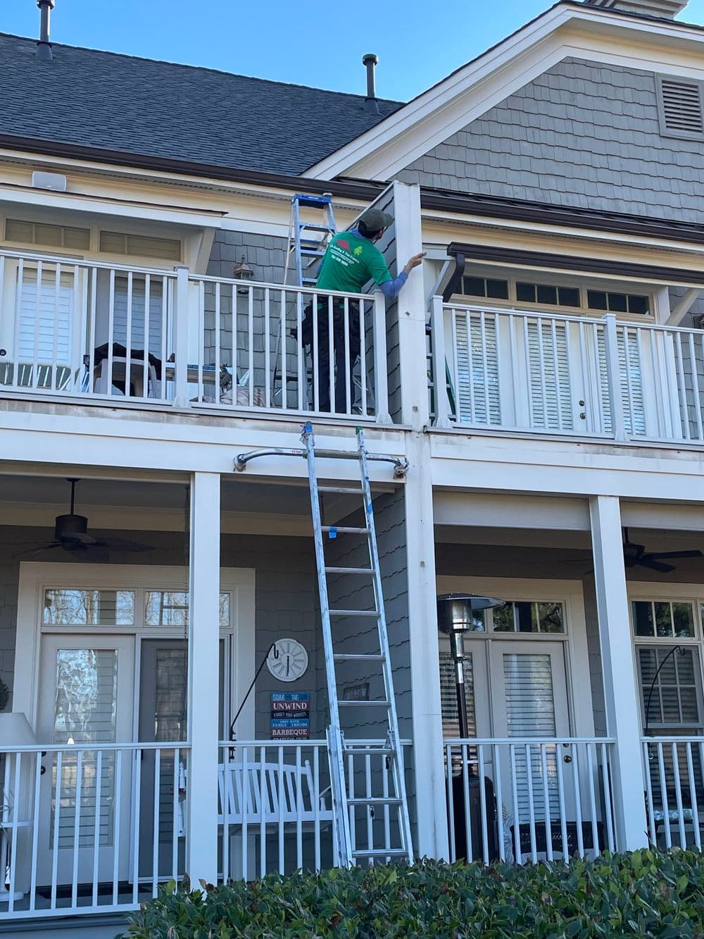 Person using a ladder to perform maintenance on a second-floor balcony of a house.