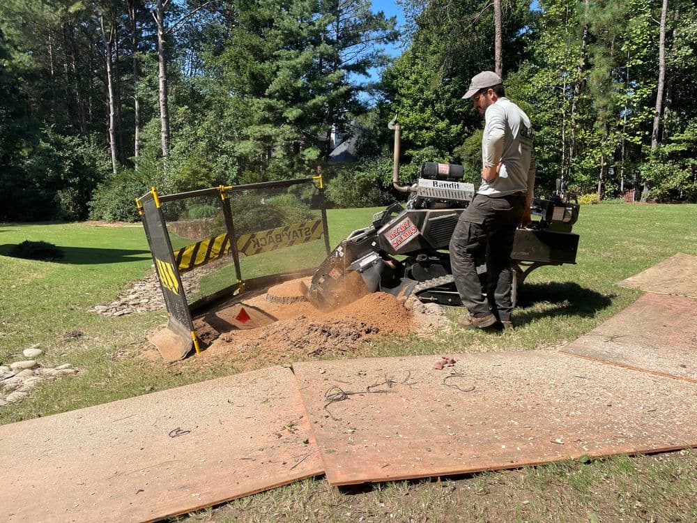 Man operating a stump grinder on a grassy area, clearing wood debris and soil.
