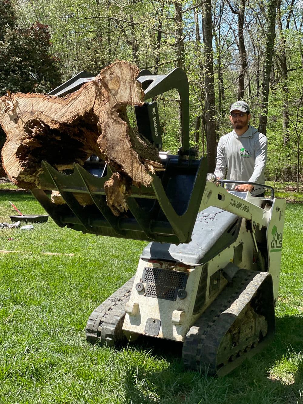 Man operating a skid steer loader to lift a large log in a green outdoor setting.