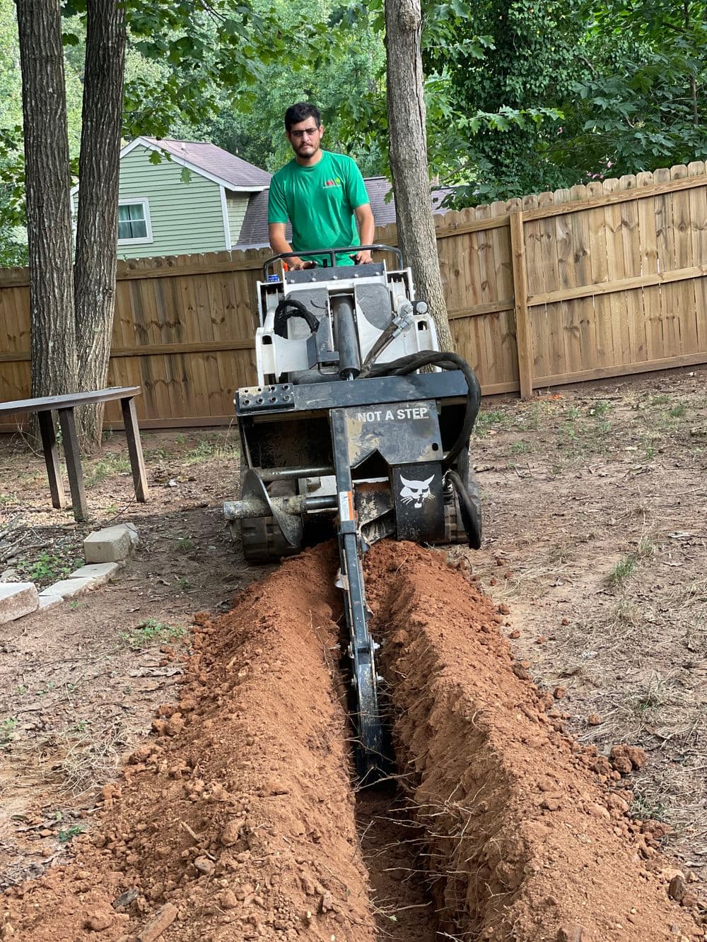 Man operating a trenching machine in a backyard, digging a narrow trench in dry soil.
