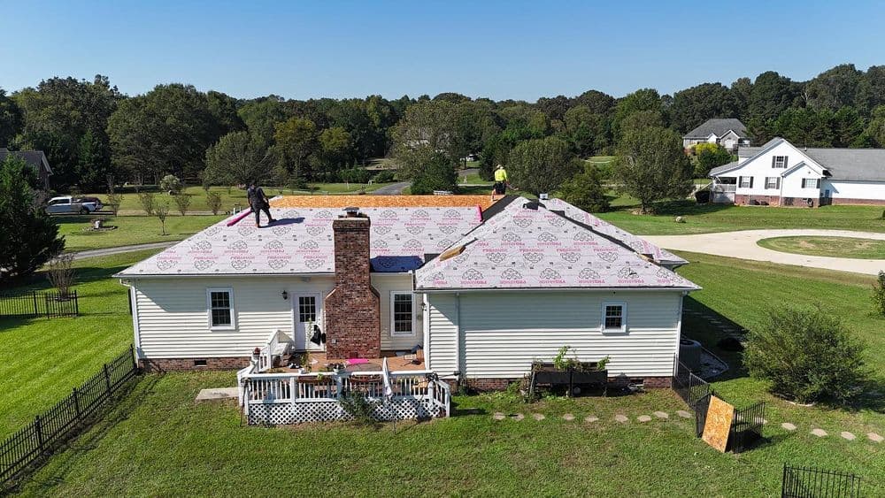 Roof installation in progress on a residential home with workers on top and green surroundings.