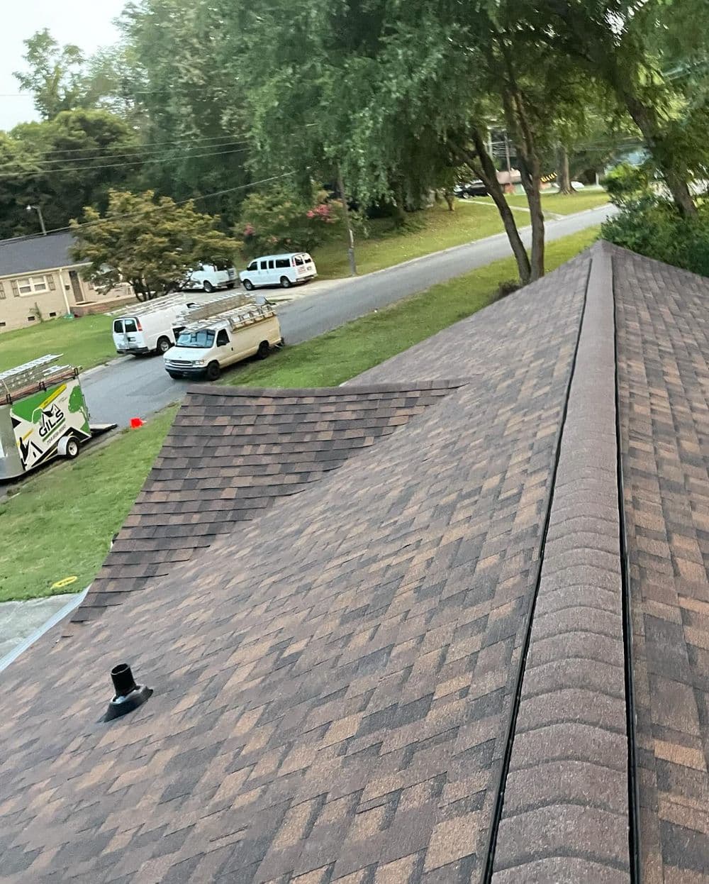 Aerial view of a brown shingle roof with nearby vehicles and green trees in the background.