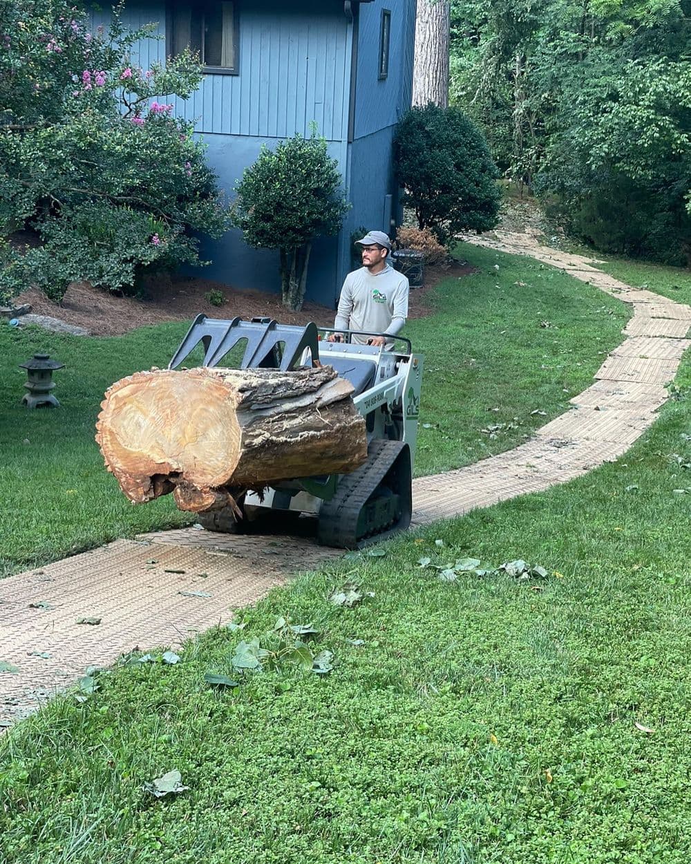 Man operating a mini loader to transport a large log on a grassy path.